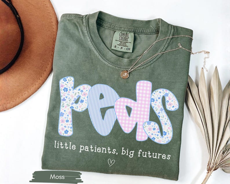 May include: Moss green t-shirt with the word "peds" in a playful font, featuring floral, striped, and gingham patterns. The text "little patients, big futures" is below the word "peds". A gold necklace and a brown hat are also in the image.