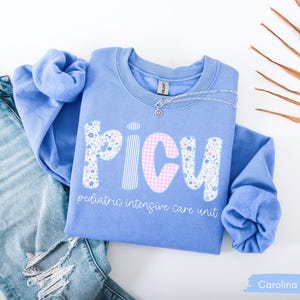 May include: Carolina blue sweatshirt with the letters "PICU" in a floral and gingham pattern, with the words "pediatric intensive care unit" below. A silver necklace and a pair of light blue jeans are next to the sweatshirt.