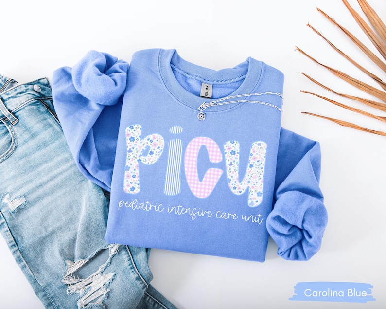 May include: Carolina blue sweatshirt with the letters "PICU" in a floral and gingham pattern, with the words "pediatric intensive care unit" below. A silver necklace and a pair of light blue jeans are next to the sweatshirt.