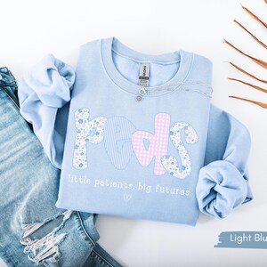 May include: Light blue sweatshirt with the word "PEDS" in floral, striped, and gingham patterns. The text "little patients, big futures" is below the word "PEDS". A silver necklace is on the sweatshirt. Light blue jeans are next to the sweatshirt.