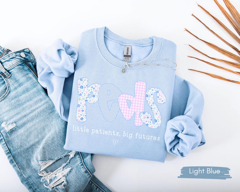 May include: Light blue sweatshirt with the word "PEDS" in floral, striped, and gingham patterns. The text "little patients, big futures" is below the word "PEDS". A silver necklace is on the sweatshirt. Light blue jeans are next to the sweatshirt.
