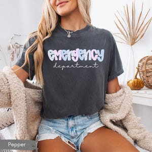 May include: Dark grey t-shirt with the words "emergency department" in a pastel, bubble font. The shirt is worn by a person with blonde hair and blue jean shorts. The shirt is a relaxed fit.