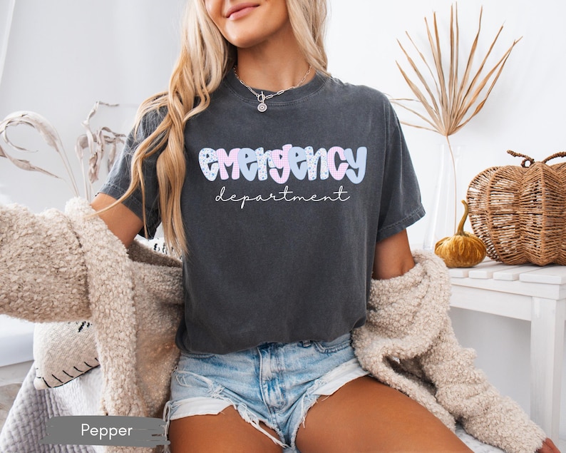 May include: Dark grey t-shirt with the words "emergency department" in a pastel, bubble font. The shirt is worn by a person with blonde hair and blue jean shorts. The shirt is a relaxed fit.