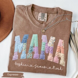 Puede incluir: Camiseta color espresso con la palabra "MAMA" en letras de patchwork. Las letras son de varios colores pastel y patrones. Debajo de la palabra "MAMA" est&aacute;n los nombres "Sophia", "James" y "Noah". Un collar plateado con un colgante est&aacute; sobre la camiseta.