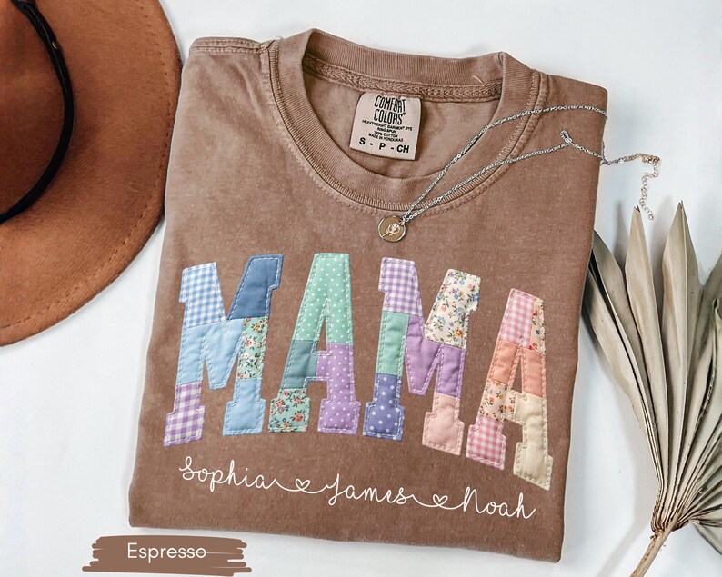 Puede incluir: Camiseta color espresso con la palabra "MAMA" en letras de patchwork. Las letras son de varios colores pastel y patrones. Debajo de la palabra "MAMA" est&aacute;n los nombres "Sophia", "James" y "Noah". Un collar plateado con un colgante est&aacute; sobre la camiseta.