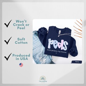 May include: Navy blue sweatshirt with the word "peds" in a floral and checkered design. The text "little patients, big futures" is below the word "peds". The sweatshirt is paired with blue jeans. The image also includes text that says "Won't Crack or Peel", "Soft Cotton", and "Produced in USA".