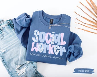 Puede incluir: Sudadera azul índigo con la frase "SOCIAL WORKER" en una fuente divertida, con las palabras "advocate. support. empower." debajo. La sudadera se muestra con vaqueros azul claro y un collar plateado.