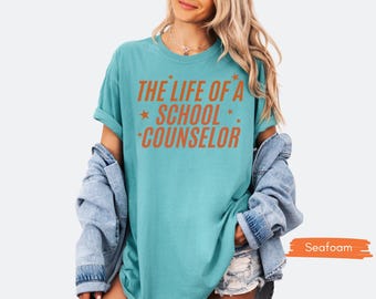 May include: Seafoam green t-shirt with the text "THE LIFE OF A SCHOOL COUNSELOR" in orange. The text is accented with orange stars. The shirt is a solid color and has rolled-up sleeves.