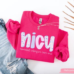 Op de afbeelding: Een felroze sweatshirt met de tekst "NICU neonatal intensive care unit". De letters zijn versierd met bloemen- en ruitpatronen. Een zilveren ketting ligt op de trui, die naast een blauwe spijkerbroek ligt.