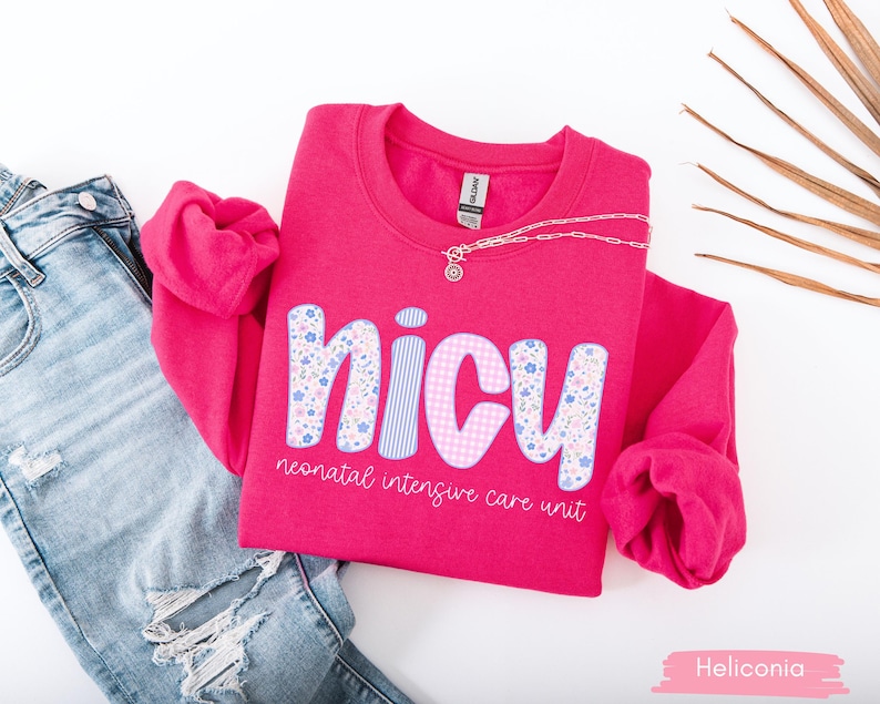 Op de afbeelding: Een felroze sweatshirt met de tekst "NICU neonatal intensive care unit". De letters zijn versierd met bloemen- en ruitpatronen. Een zilveren ketting ligt op de trui, die naast een blauwe spijkerbroek ligt.