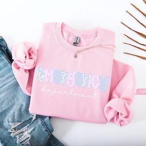 May include: A light pink sweatshirt with the words "EMERGENCY department" in a playful font. The word "EMERGENCY" is in a blue floral pattern and light blue letters. The sweatshirt is paired with ripped blue jeans and a delicate necklace.