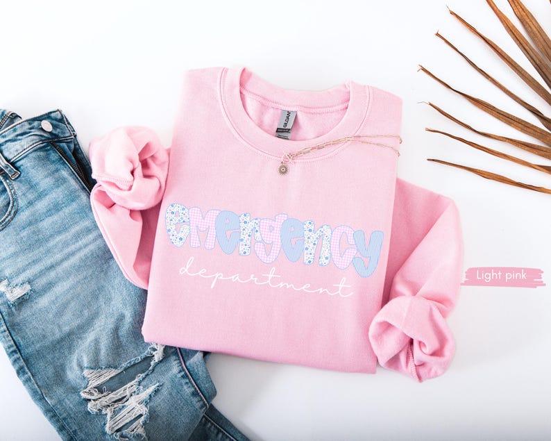 May include: A light pink sweatshirt with the words "EMERGENCY department" in a playful font. The word "EMERGENCY" is in a blue floral pattern and light blue letters. The sweatshirt is paired with ripped blue jeans and a delicate necklace.