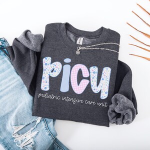 May include: A dark gray sweatshirt with the letters "PICU" in floral and striped patterns. The text "pediatric intensive care unit" is below the letters. A silver necklace and blue jeans are also in the image.