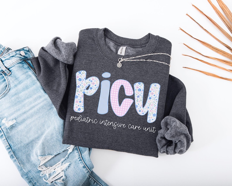 May include: A dark gray sweatshirt with the letters "PICU" in floral and striped patterns. The text "pediatric intensive care unit" is below the letters. A silver necklace and blue jeans are also in the image.