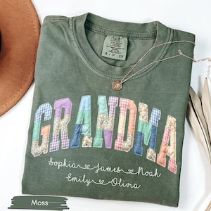 May include: A moss green t-shirt with the word "GRANDMA" in colorful patchwork letters. Below the word are the names Sophia, James, Noah, Emily, and Olivia. A gold necklace rests on the shirt.