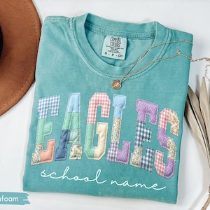 May include: Seafoam green t-shirt with the word "EAGLES" in patchwork letters. The letters are in various patterns and colors. The words "school name" are written in cursive below the letters. A gold necklace is draped across the shirt.