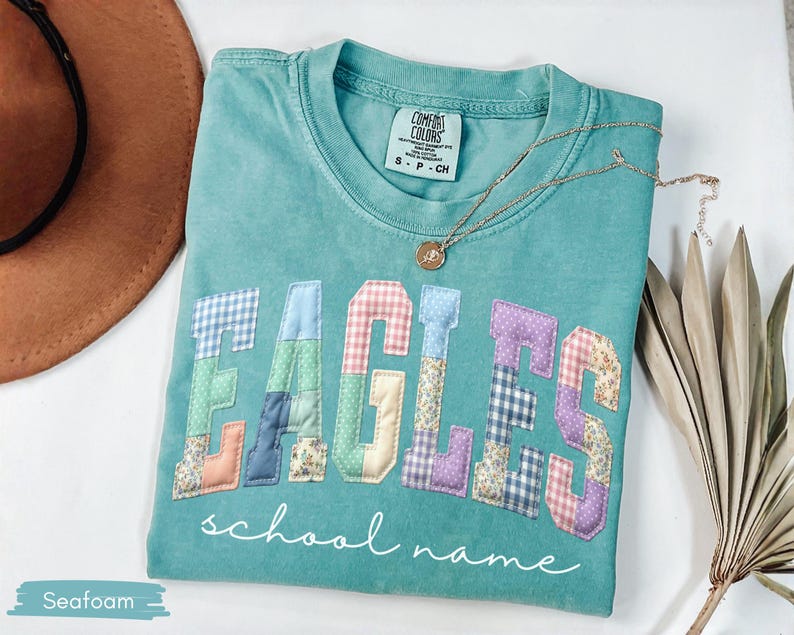 May include: Seafoam green t-shirt with the word "EAGLES" in patchwork letters. The letters are in various patterns and colors. The words "school name" are written in cursive below the letters. A gold necklace is draped across the shirt.