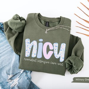 Op de afbeelding: Militairgroen sweatshirt met het woord "NICU" in een bloemen-, ruit- en streepontwerp. De tekst "neonatal intensive care unit" staat onder het woord "NICU". Een zilveren ketting ligt op het sweatshirt. Het sweatshirt ligt naast een spijkerbroek.