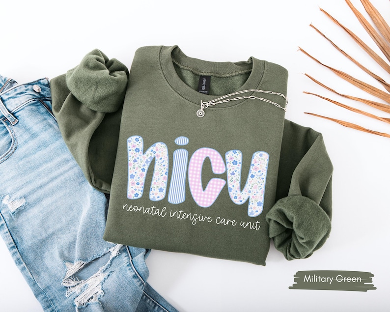 Op de afbeelding: Militairgroen sweatshirt met het woord "NICU" in een bloemen-, ruit- en streepontwerp. De tekst "neonatal intensive care unit" staat onder het woord "NICU". Een zilveren ketting ligt op het sweatshirt. Het sweatshirt ligt naast een spijkerbroek.