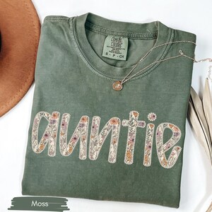 May include: A moss green t-shirt with the word "Auntie" embroidered in a floral design. A gold necklace with a small pendant rests on the shirt. A brown hat and dried plant are in the background.