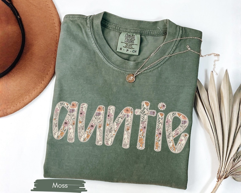 May include: A moss green t-shirt with the word "Auntie" embroidered in a floral design. A gold necklace with a small pendant rests on the shirt. A brown hat and dried plant are in the background.