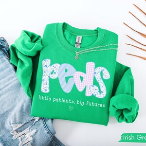 May include: A bright green sweatshirt with the word "Peds" in a mix of floral, striped, and gingham patterns. The text "little patients, big futures" is below the word "Peds". A silver necklace and blue jeans are next to the sweatshirt.