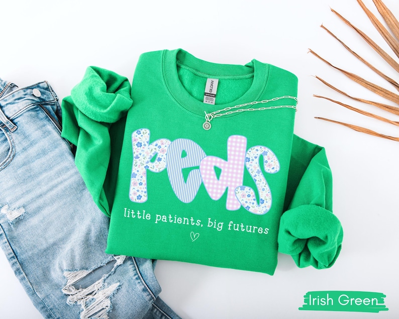 May include: A bright green sweatshirt with the word "Peds" in a mix of floral, striped, and gingham patterns. The text "little patients, big futures" is below the word "Peds". A silver necklace and blue jeans are next to the sweatshirt.