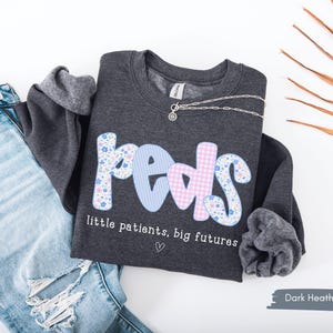 May include: Dark heather gray sweatshirt with the word "peds" in a floral, striped, and gingham pattern. The text "little patients, big futures" is below the word "peds". A silver necklace is draped across the sweatshirt. Light blue jeans are next to the sweatshirt.