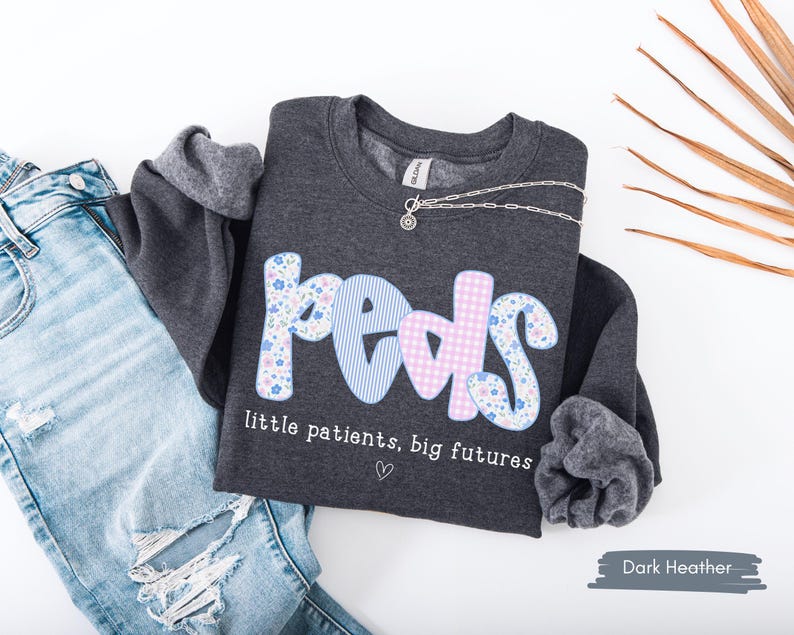 May include: Dark heather gray sweatshirt with the word "peds" in a floral, striped, and gingham pattern. The text "little patients, big futures" is below the word "peds". A silver necklace is draped across the sweatshirt. Light blue jeans are next to the sweatshirt.