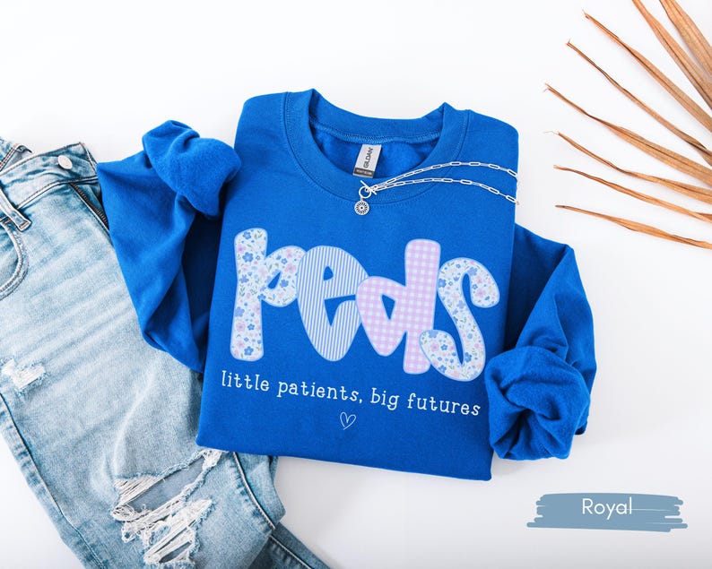 May include: Royal blue sweatshirt with the word "Peds" in a patchwork design. The letters are decorated with floral, striped, and gingham patterns. The text "little patients, big futures" is below the word "Peds". A silver necklace is on the sweatshirt.