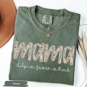 Puede incluir: Camiseta verde musgo con la palabra "MAMA" bordada con flores. Debajo, los nombres "Lily", "James" y "Noah". Un collar dorado con un colgante descansa sobre la camiseta. Un sombrero marr&oacute;n y una planta seca est&aacute;n en el fondo.
