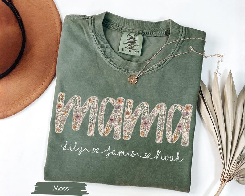 Puede incluir: Camiseta verde musgo con la palabra "MAMA" bordada con flores. Debajo, los nombres "Lily", "James" y "Noah". Un collar dorado con un colgante descansa sobre la camiseta. Un sombrero marr&oacute;n y una planta seca est&aacute;n en el fondo.