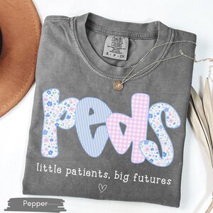 May include: A gray t-shirt with the word "peds" in a floral, gingham, and striped design. The text "little patients, big futures" is below the word "peds". A gold necklace is on the shirt.