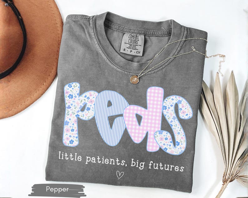 May include: A gray t-shirt with the word "peds" in a floral, gingham, and striped design. The text "little patients, big futures" is below the word "peds". A gold necklace is on the shirt.
