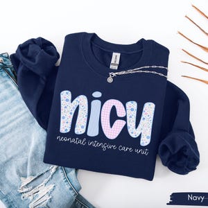 Op de afbeelding: Marineblauw sweatshirt met het woord "NICU" in een speels lettertype. De letters zijn versierd met bloemen-, streep- en ruitpatronen. De tekst "neonatal intensive care unit" staat eronder. Een zilveren ketting ligt op de trui.