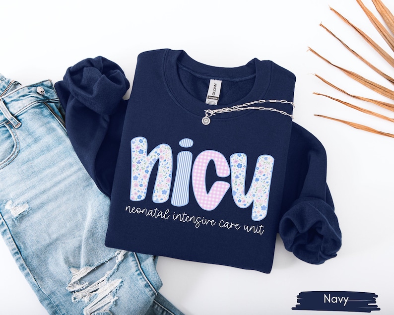Op de afbeelding: Marineblauw sweatshirt met het woord "NICU" in een speels lettertype. De letters zijn versierd met bloemen-, streep- en ruitpatronen. De tekst "neonatal intensive care unit" staat eronder. Een zilveren ketting ligt op de trui.