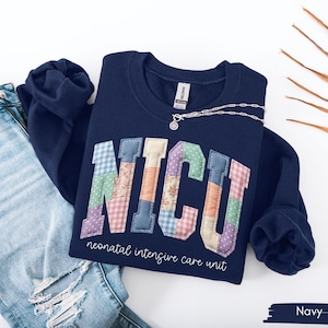 May include: Navy blue sweatshirt with the letters "NICU" in a patchwork design. The words "neonatal intensive care unit" are printed below the letters. A silver necklace with a small pendant rests on the sweatshirt. Light blue jeans are visible.