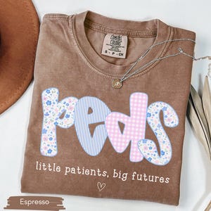 May include: Espresso-colored t-shirt with the word "PEDS" in a floral, striped, and gingham design. The text "little patients, big futures" is below the word "PEDS". A silver necklace and a brown hat are also visible.