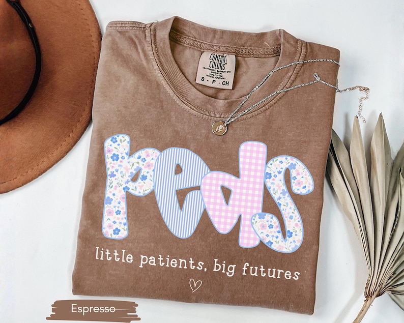 May include: Espresso-colored t-shirt with the word "PEDS" in a floral, striped, and gingham design. The text "little patients, big futures" is below the word "PEDS". A silver necklace and a brown hat are also visible.