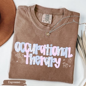 Op de afbeelding: Espresso-kleurig T-shirt met de tekst "Occupational Therapy" in een speels, kleurrijk lettertype. Het ontwerp bevat puzzelstukjes, scharen en andere pictogrammen die gerelateerd zijn aan ergotherapie. Een zilveren ketting met een hanger ligt over het shirt.