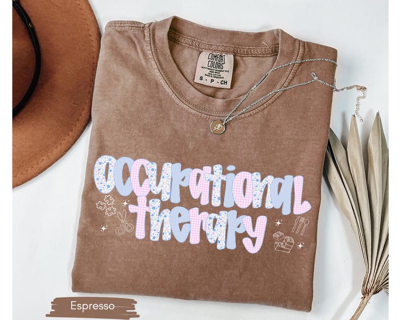 Op de afbeelding: Espresso-kleurig T-shirt met de tekst "Occupational Therapy" in een speels, kleurrijk lettertype. Het ontwerp bevat puzzelstukjes, scharen en andere pictogrammen die gerelateerd zijn aan ergotherapie. Een zilveren ketting met een hanger ligt over het shirt.