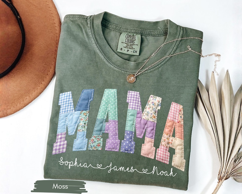 Puede incluir: Camiseta verde musgo con la palabra "MAMA" en letras de patchwork. Las letras tienen varios patrones y colores. Debajo de la palabra "MAMA" est&aacute;n los nombres "Sophia", "James" y "Noah". Un collar dorado est&aacute; sobre la camiseta.