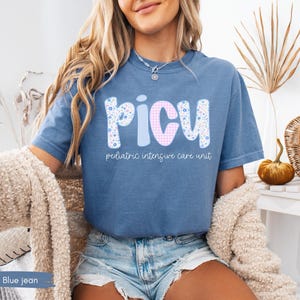 Puede incluir: Camiseta azul vaquero con las letras "PICU" en un dise&ntilde;o floral, a rayas y a cuadros. Las palabras "pediatric intensive care unit" est&aacute;n impresas debajo. La camiseta est&aacute; hecha de un material suave.