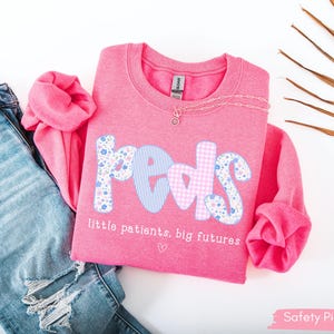 May include: A pink sweatshirt with the word "PEDS" in a mix of floral, striped, and gingham patterns. The text "little patients, big futures" is below the word. A silver necklace and ripped blue jeans are also in the image.