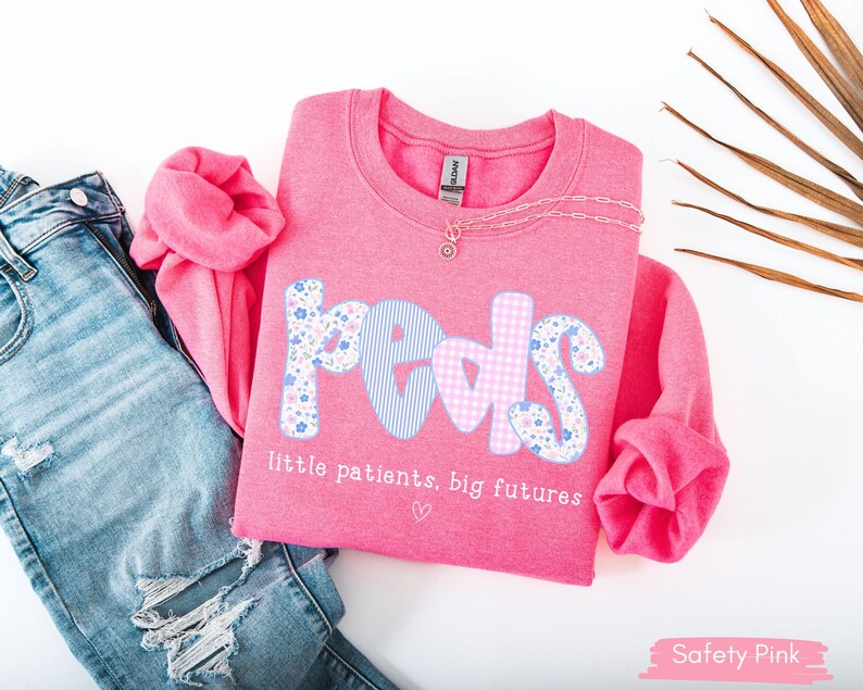 May include: A pink sweatshirt with the word "PEDS" in a mix of floral, striped, and gingham patterns. The text "little patients, big futures" is below the word. A silver necklace and ripped blue jeans are also in the image.
