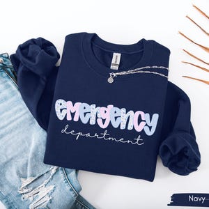 May include: Navy blue sweatshirt with the words "Emergency department" in a playful font. The word "Emergency" is in a mix of light blue, pink, and patterned letters. A silver necklace with a pendant is draped on the sweatshirt.