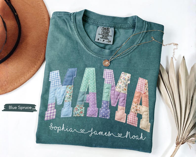 Puede incluir: Una camiseta Comfort Colors verde azulado con la palabra "MAMA" en letras de patchwork. Las letras est&aacute;n hechas de varias telas estampadas. Debajo de la palabra "MAMA" est&aacute;n los nombres "Sophia", "James" y "Noah". Tambi&eacute;n se ven un collar dorado y un sombrero marr&oacute;n.