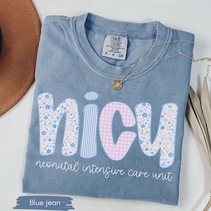 Peut inclure: T-shirt bleu jean avec le mot "NICU" dans un motif floral et &agrave; motifs. Le texte "neonatal intensive care unit" est en dessous du mot "NICU". Un collier en or est drap&eacute; sur la chemise.
