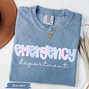 May include: Blue jean Comfort Colors t-shirt with the words "emergency department" in a playful font. The word "emergency" is in a mix of pink gingham and floral patterns. A gold necklace is draped across the shirt.