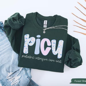 May include: Forest green sweatshirt with the letters "PICU" in a floral and gingham pattern. The words "pediatric intensive care unit" are printed below the letters. A silver necklace is draped across the sweatshirt, which is laid on top of a pair of blue jeans.